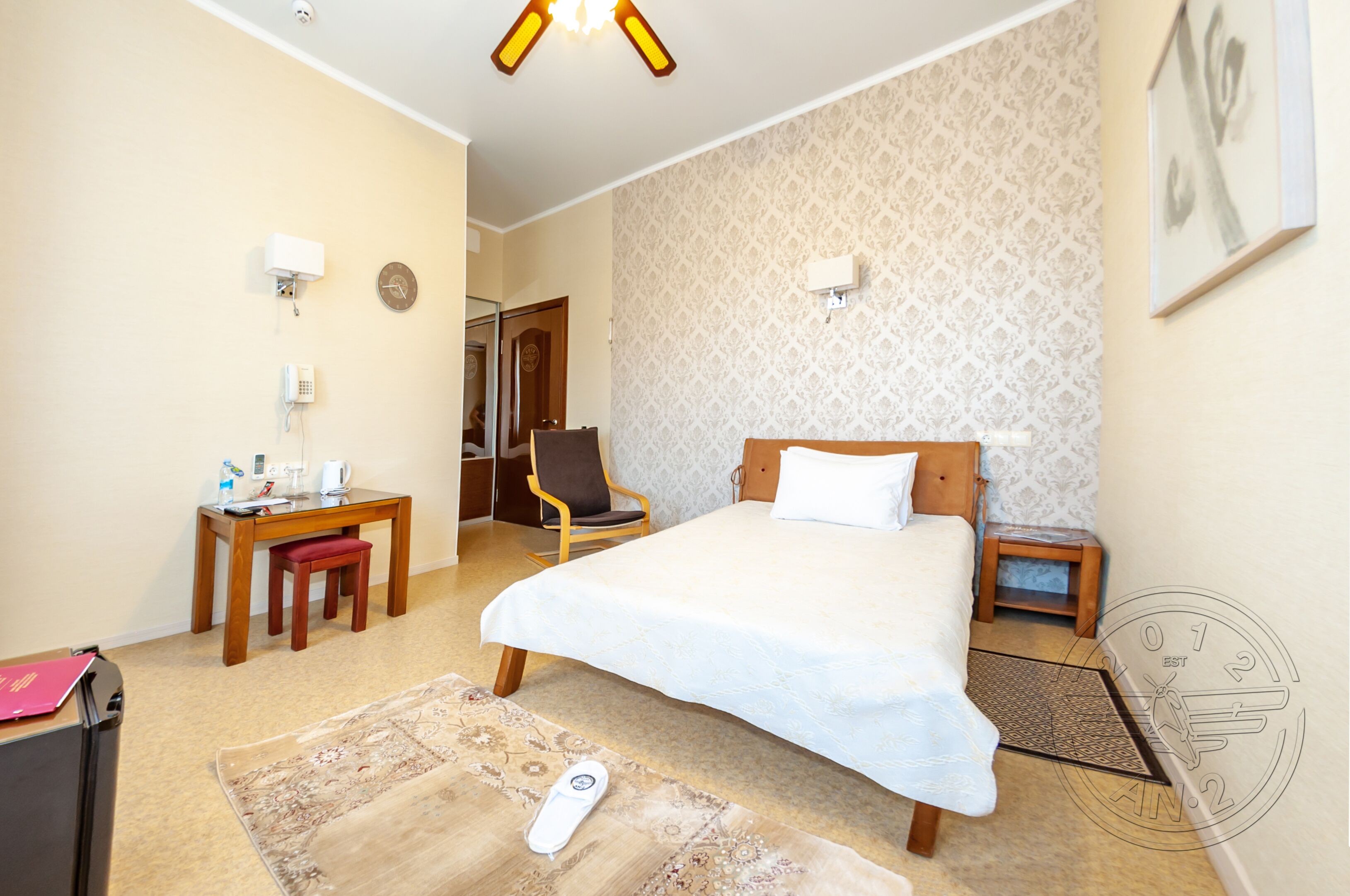 A hotel room with a bed, a desk, a chair, a small table, a nightstand, a wall clock, a thermostat, and a door leading to another room.