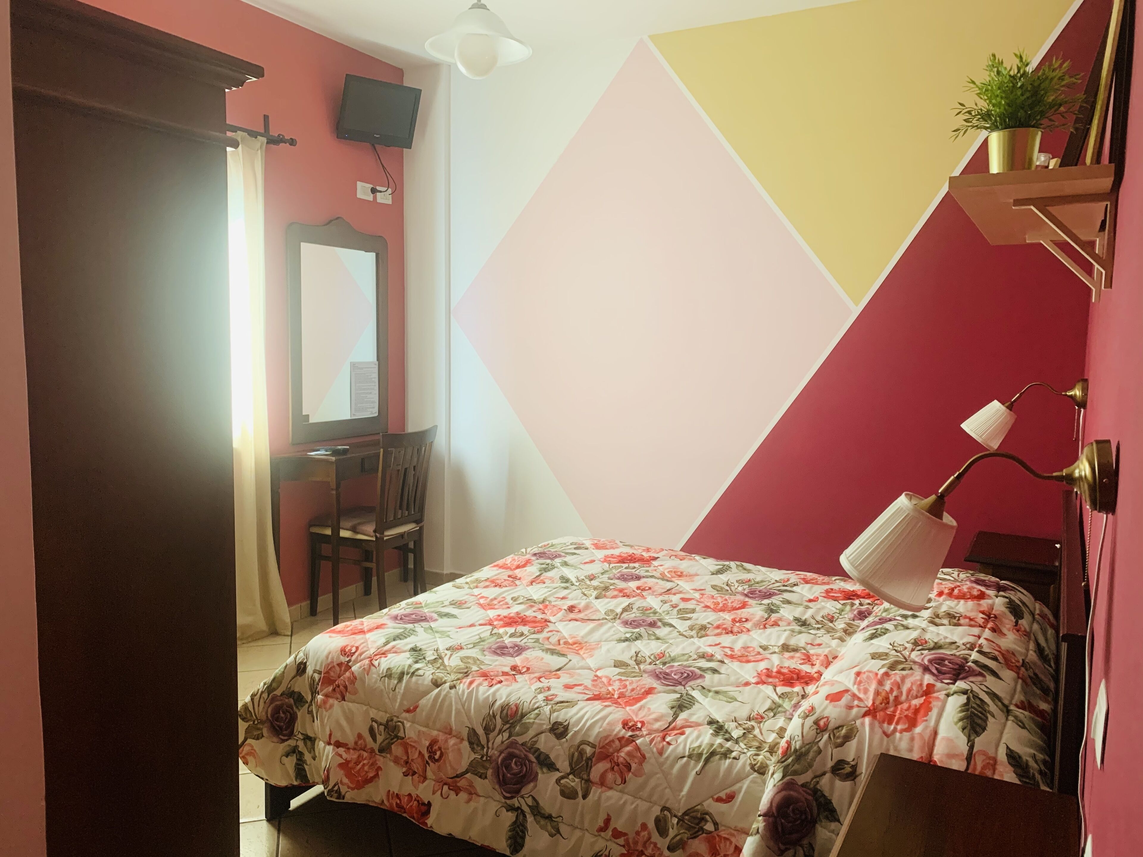 A bedroom with a floral bedspread, a desk with a chair, a TV mounted on the wall, and a bedside lamp.