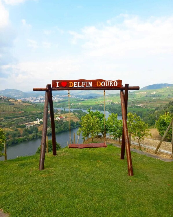 A swing set with a sign that reads "I ♥ DELFIM DOURO" overlooking a scenic landscape with a river and hills.