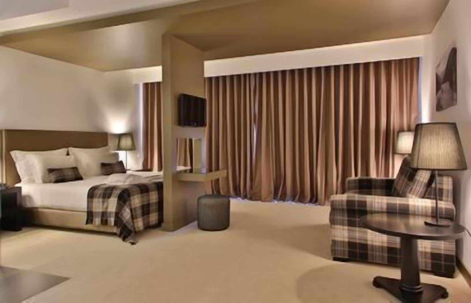 A hotel room with a bed, a chair, a small table, a lamp, and a curtain.