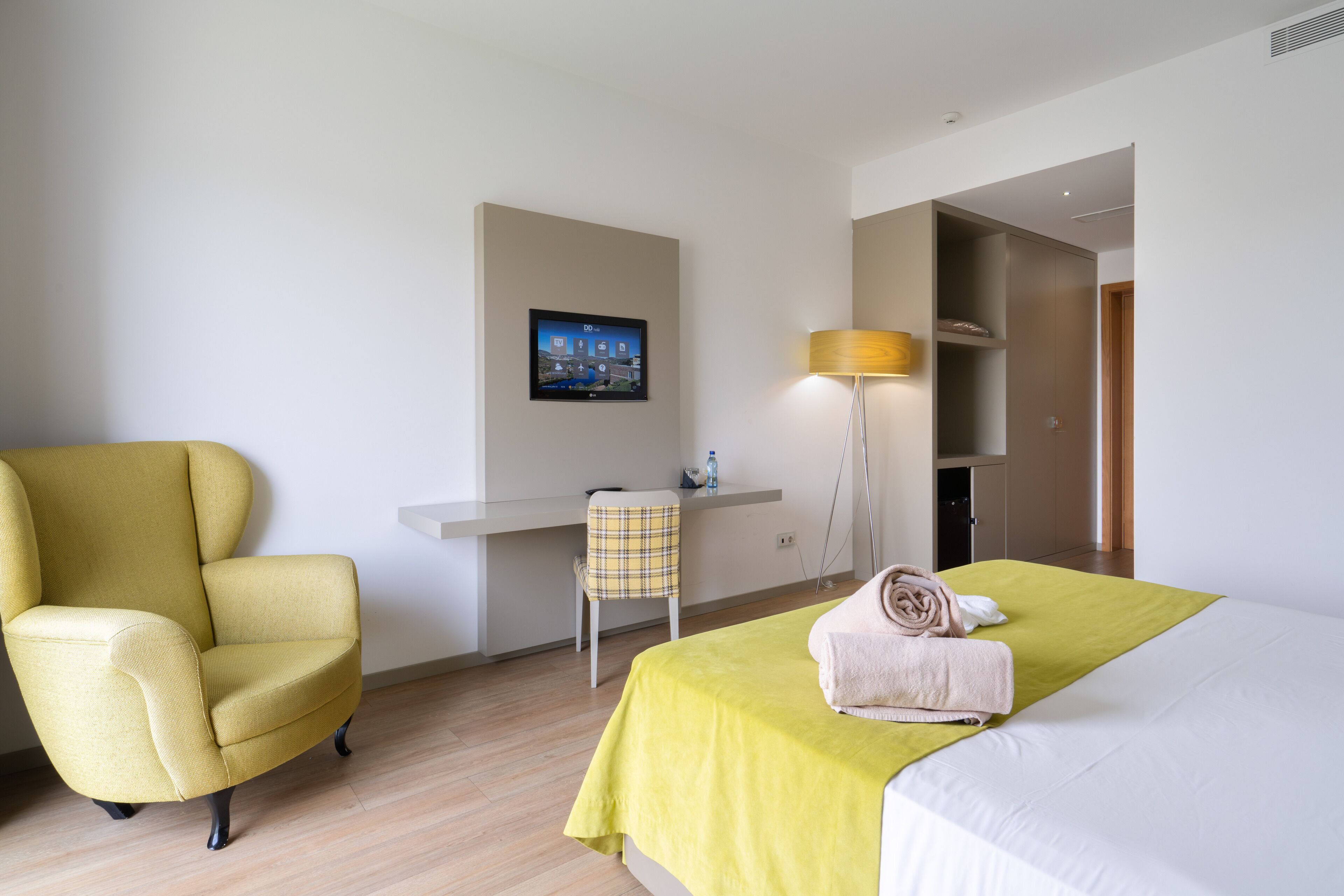 A hotel room with a yellow armchair, a desk with a chair, a bed with a green bedspread, and a lamp.