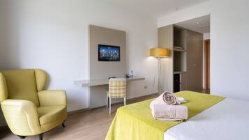 A hotel room with a yellow armchair, a desk with a chair, a bed with a green bedspread, and a lamp.