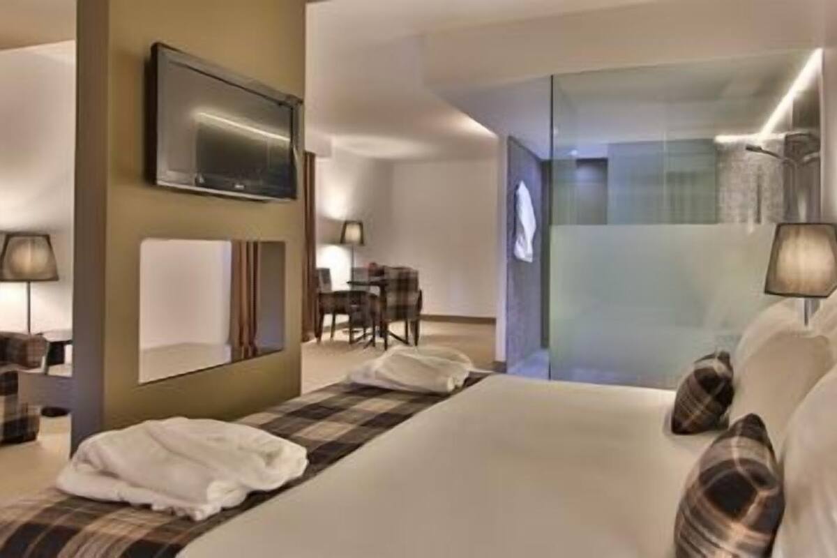 A modern hotel room with a large bed, a flat-screen TV mounted on the wall, a desk with a chair, and a glass partition separating the room from the hallway.