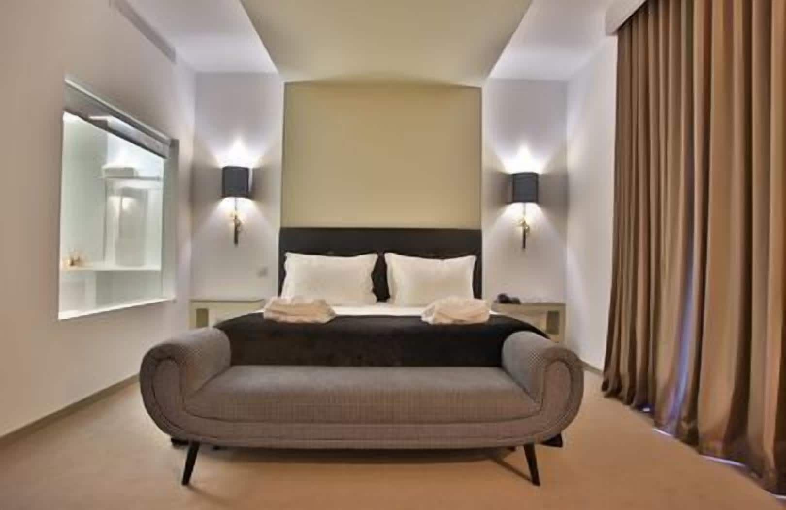 Une chambre d’hôtel moderne avec un grand lit, deux lampes de chevet, un canapé, un grand miroir et une vue sur la ville.