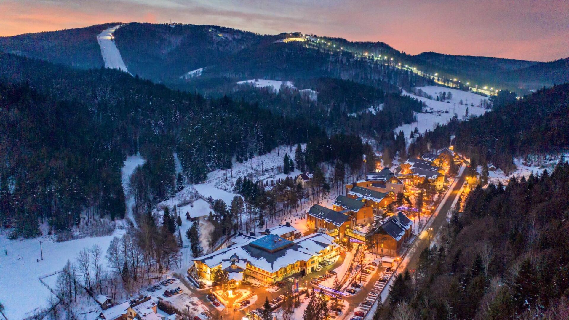 Vila coberta de neve com edifícios iluminados e uma pista de esqui ao fundo.