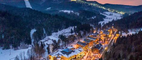 A snow-covered village with illuminated buildings and a ski slope in the background.