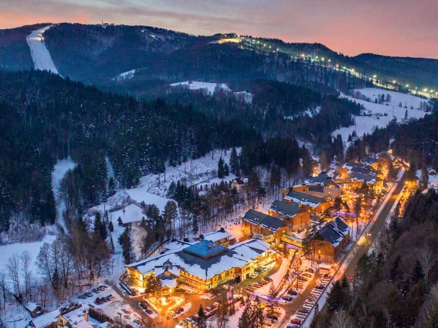 Vila coberta de neve com edifícios iluminados e uma pista de esqui ao fundo.