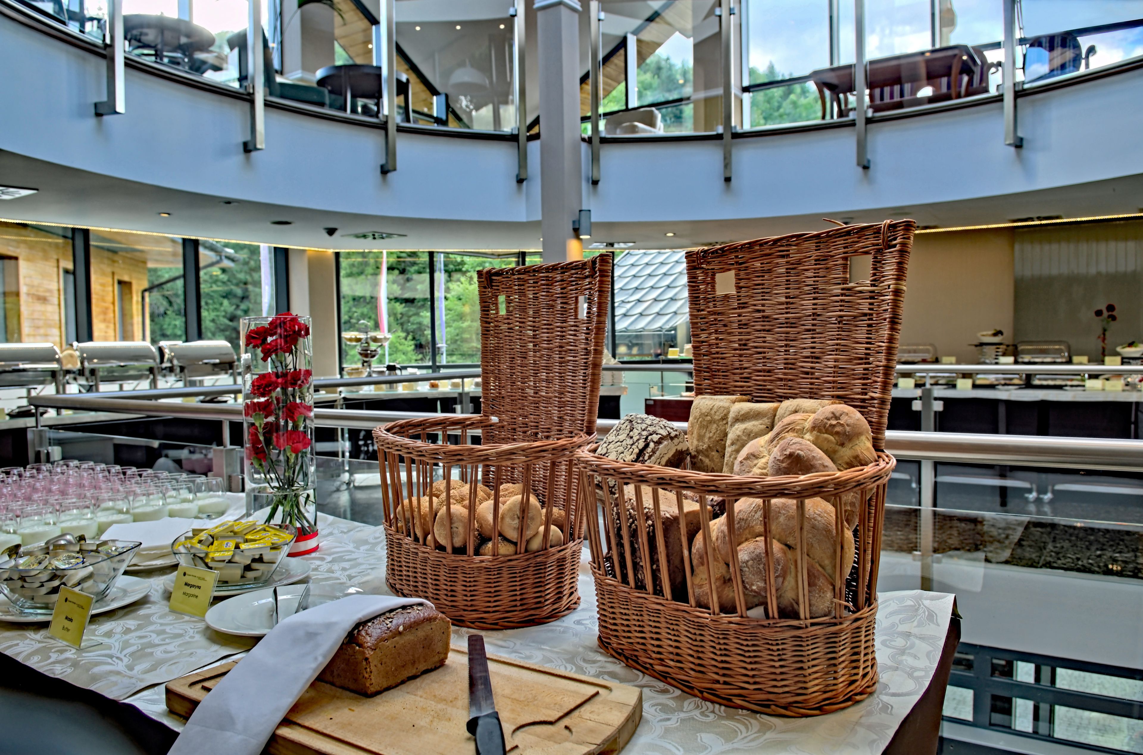 Un buffet est installé avec des paniers en osier remplis de pain et d’autres aliments, un vase en verre contenant des fleurs rouges et une planche à découper avec un couteau, le tout sur une table dans un bâtiment moderne doté de grandes fenêtres.