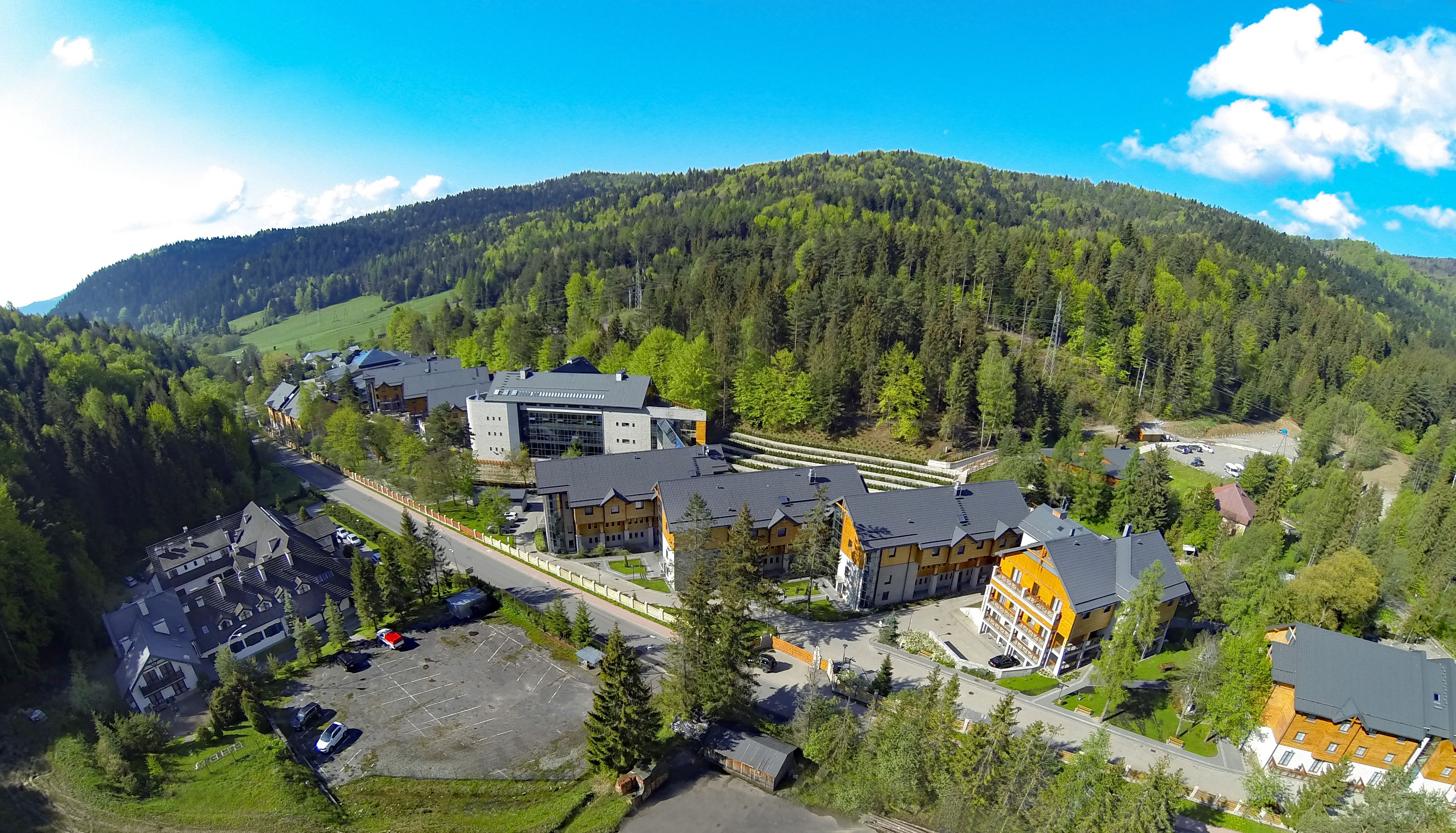An aerial view of a resort with modern buildings, a parking area, and a forested mountain backdrop.