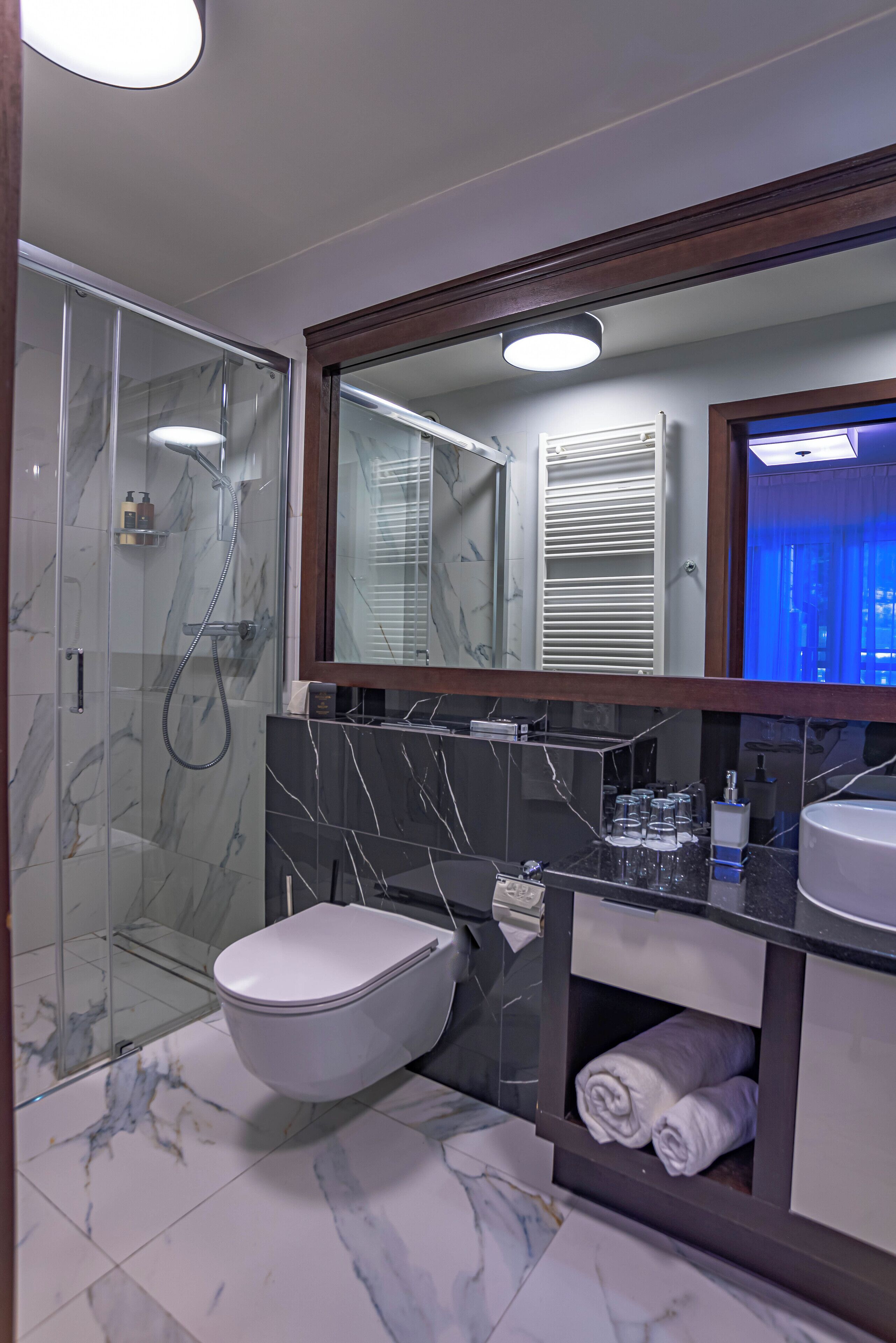 A modern bathroom with a glass-enclosed shower, a white toilet, a marble-tiled floor, a large mirror, and a built-in vanity with a sink and towel storage.