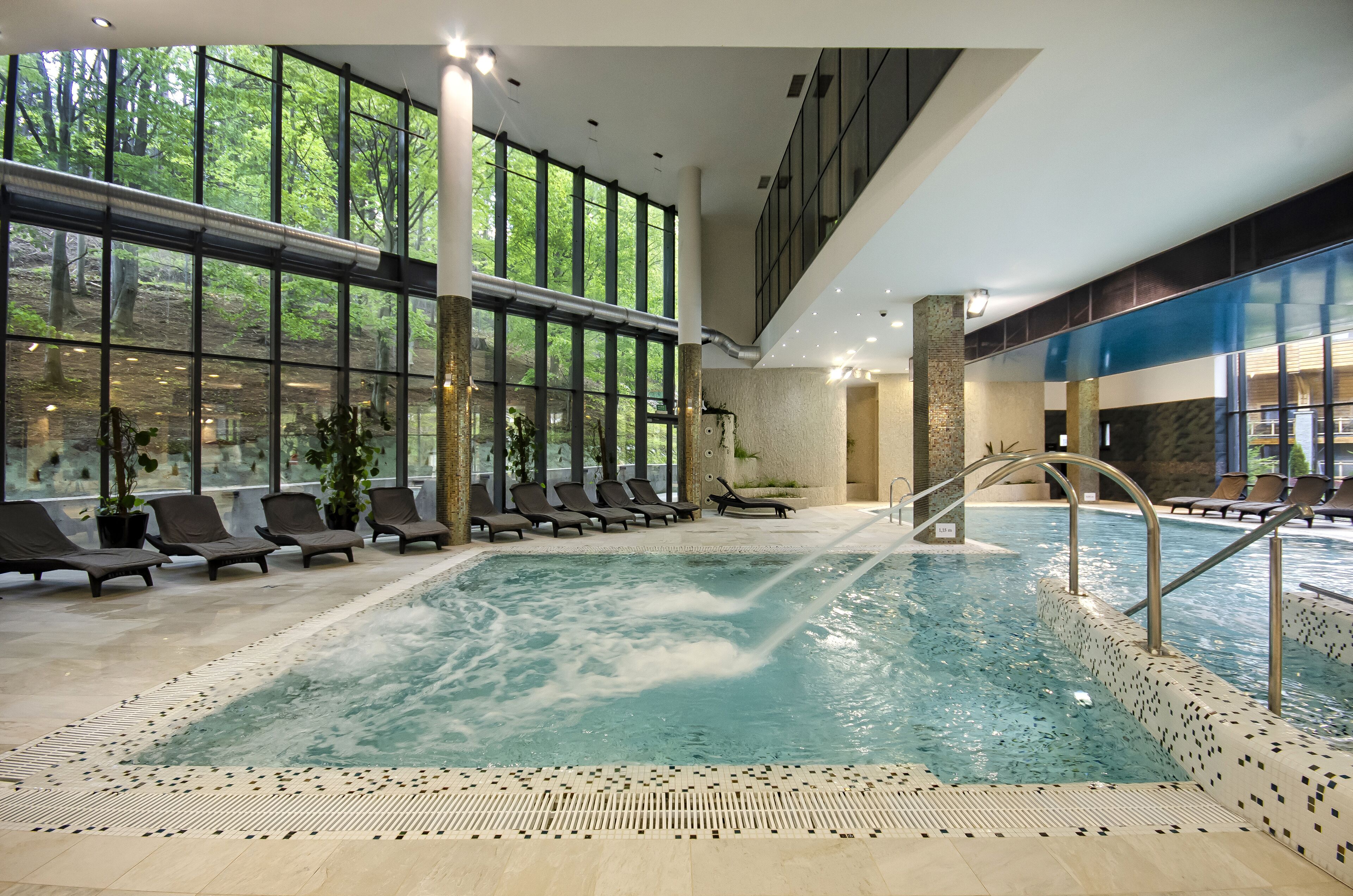 Une piscine intérieure offrant une vue dégagée sur les arbres grâce à de grandes fenêtres, des chaises longues et un sol carrelé.