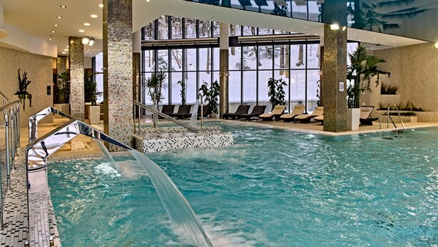 Una piscina interior con fuente de agua cristalina, sillas de descanso y ventanas amplias con vistas a árboles.