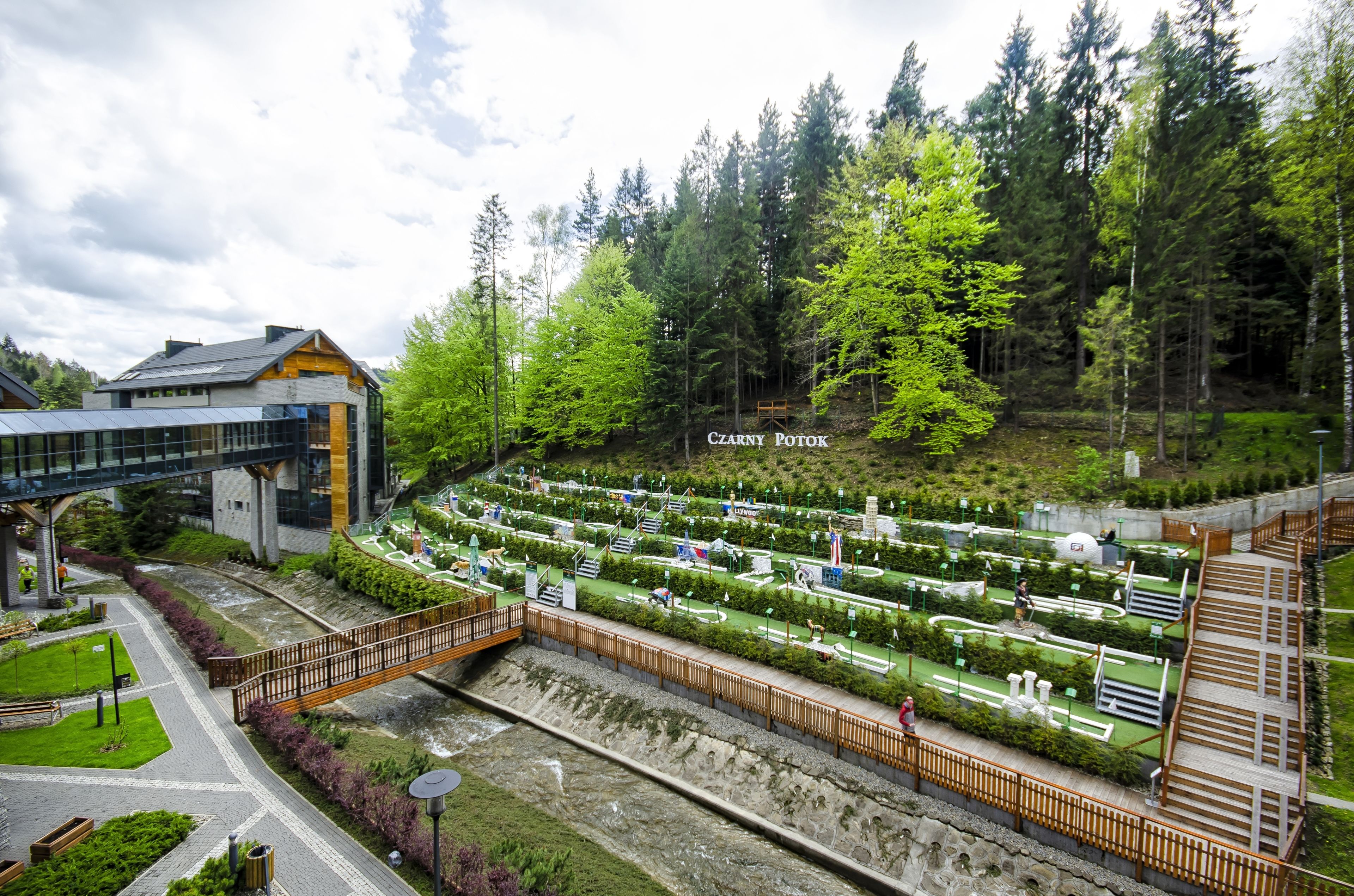A modern building with a glass facade, a wooden bridge, and a well-maintained garden with a variety of plants and a small pond.