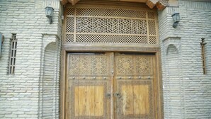 Una gran puerta de madera con elaborados grabados y tachuelas de metal, ubicada en un edificio de piedra con ventanas en arco y celosías decorativas.