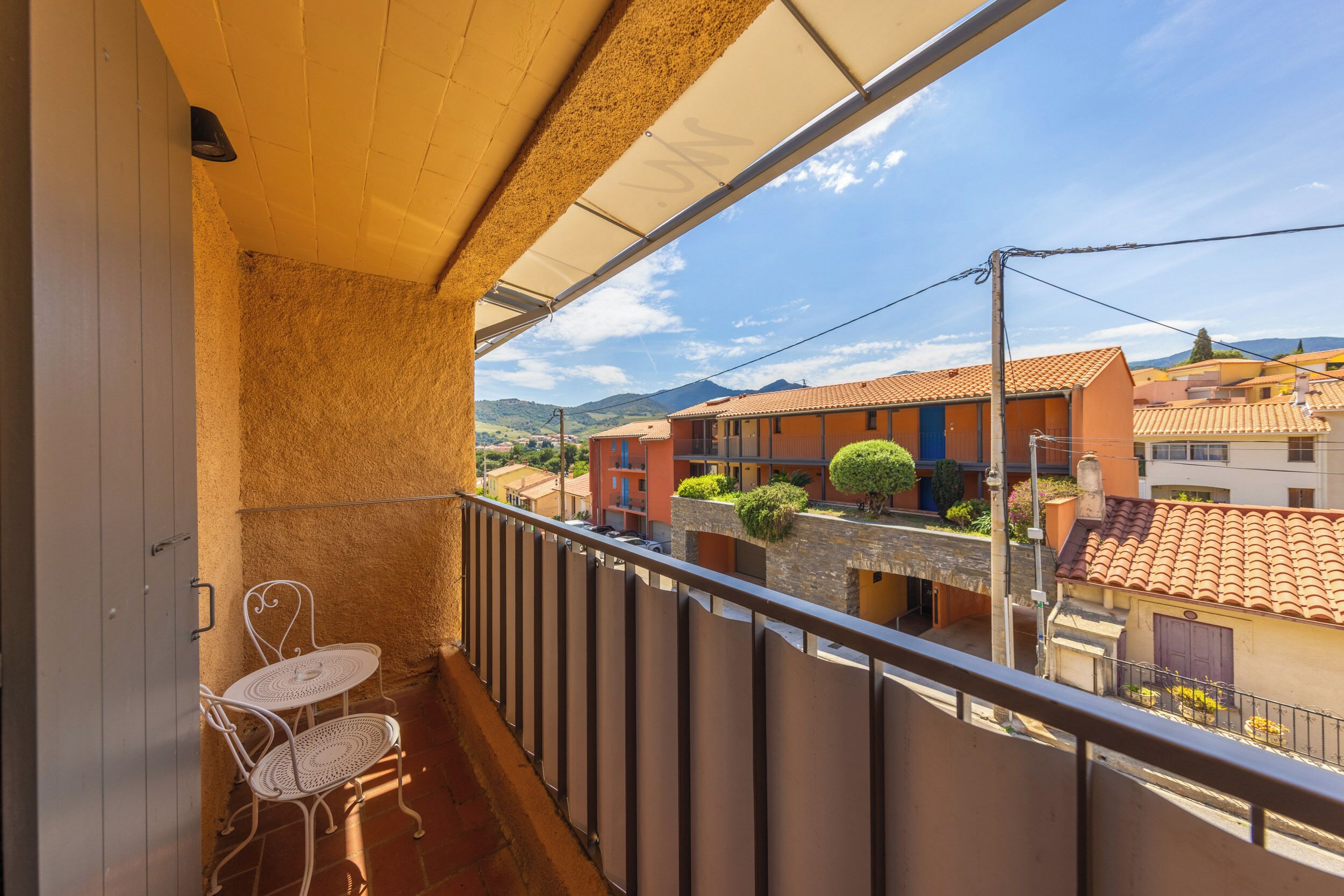A balcony with a white metal table and chairs, a view of neighboring buildings, and a clear sky.