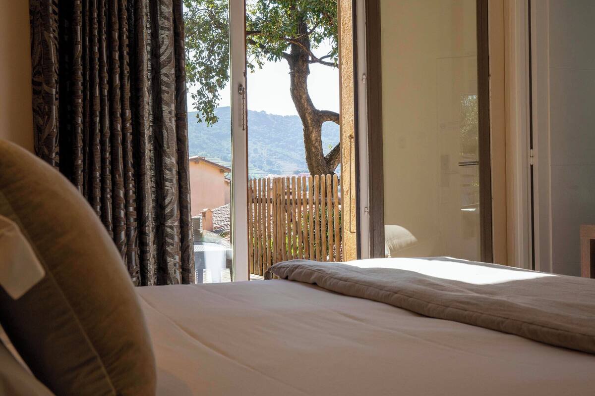 A bedroom with a bed, curtains, and a view of a tree and mountains through a glass door.