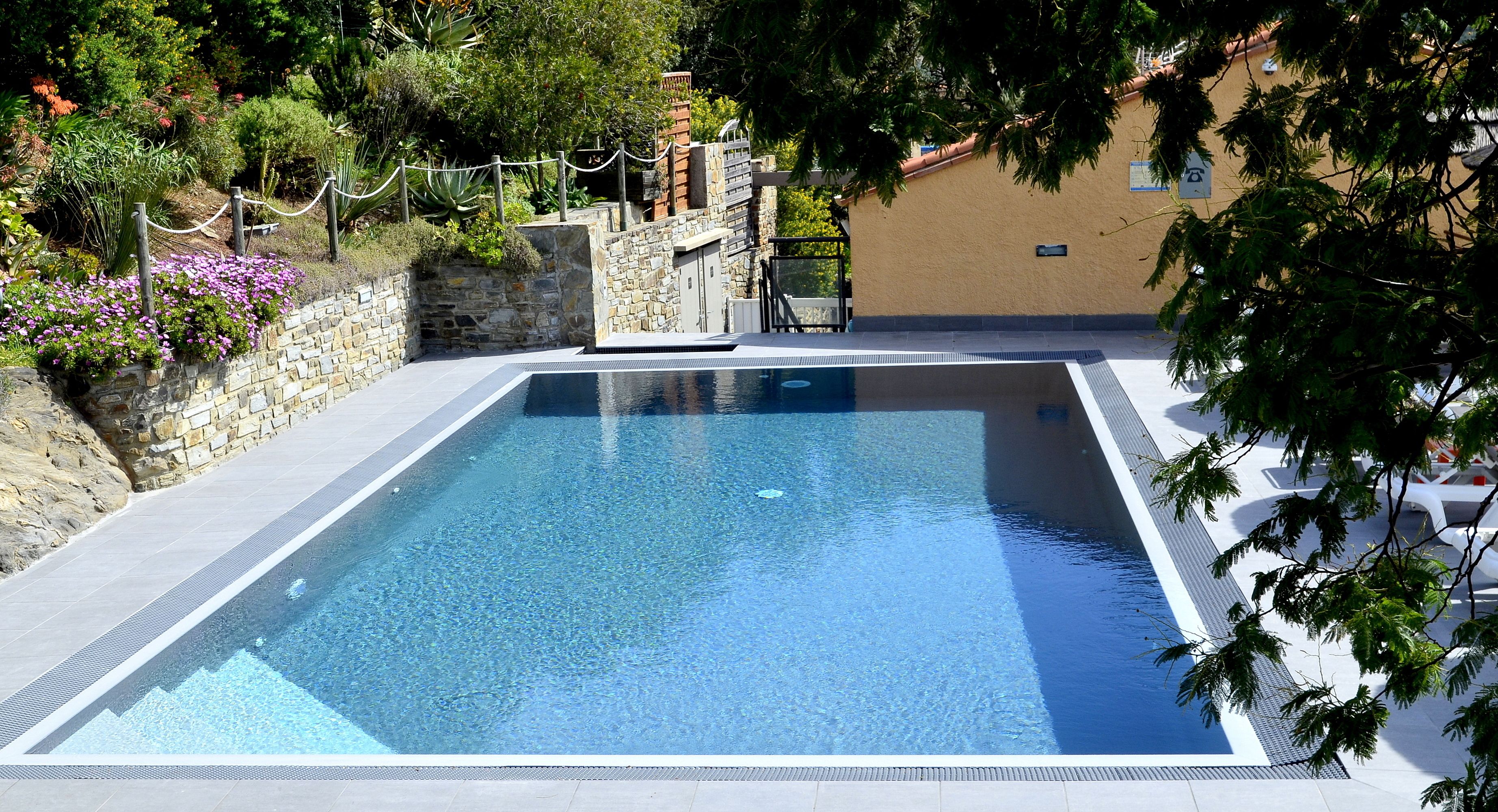Ein rechteckiger Swimmingpool mit klarem, blauem Wasser, umgeben von einer Steinmauer und einem Holzzaun.
