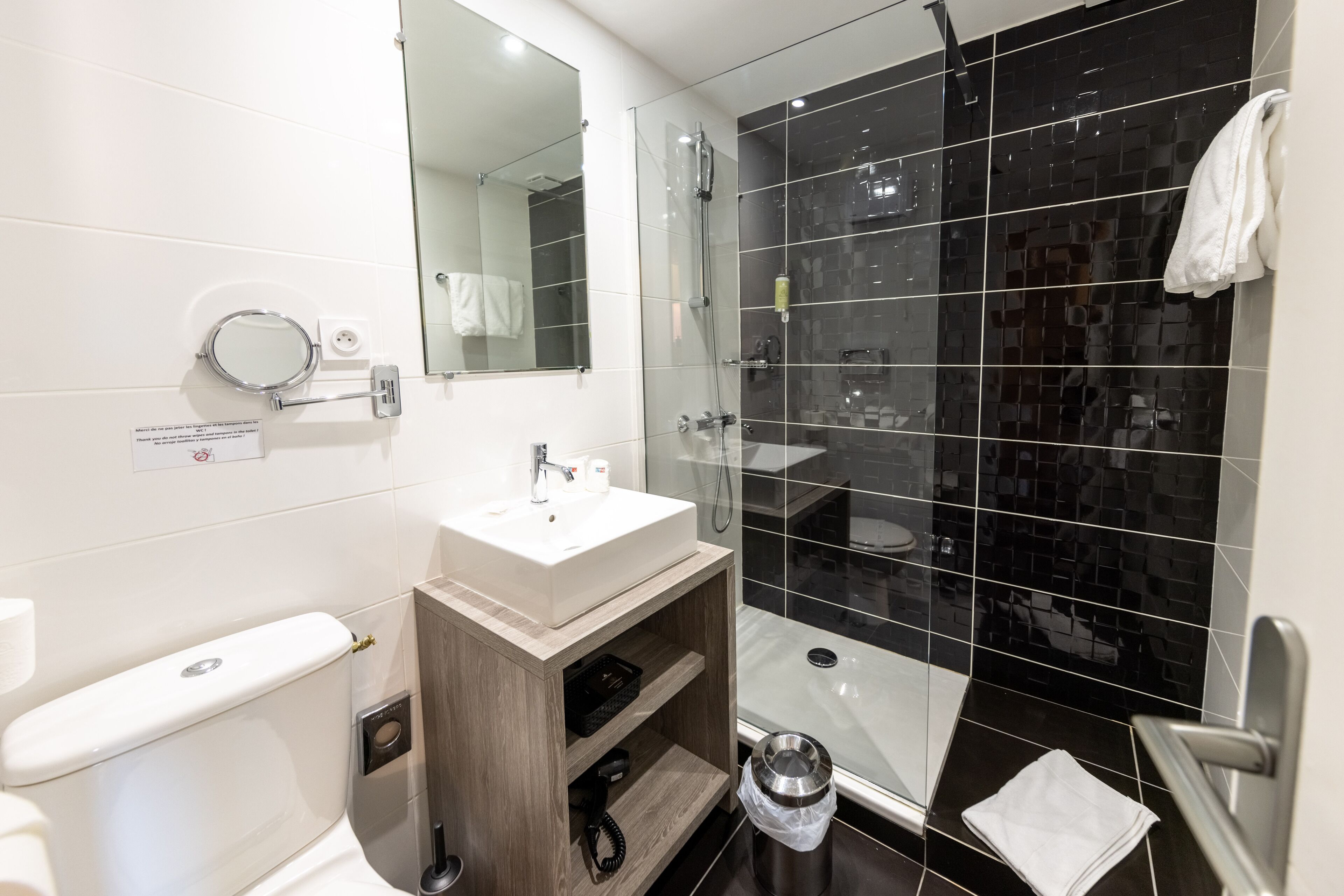 Une salle de bain moderne avec un lavabo blanc, un miroir et des toilettes. Elle est dotée de murs recouverts de carreaux noirs, d’une douche en verre et d’un meuble-lavabo en bois.