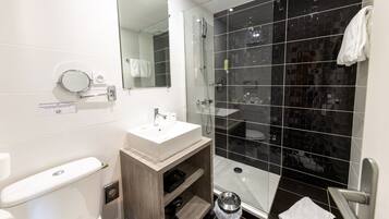 Une salle de bain moderne avec un lavabo blanc, un miroir et des toilettes. Elle est dotée de murs recouverts de carreaux noirs, d’une douche en verre et d’un meuble-lavabo en bois.