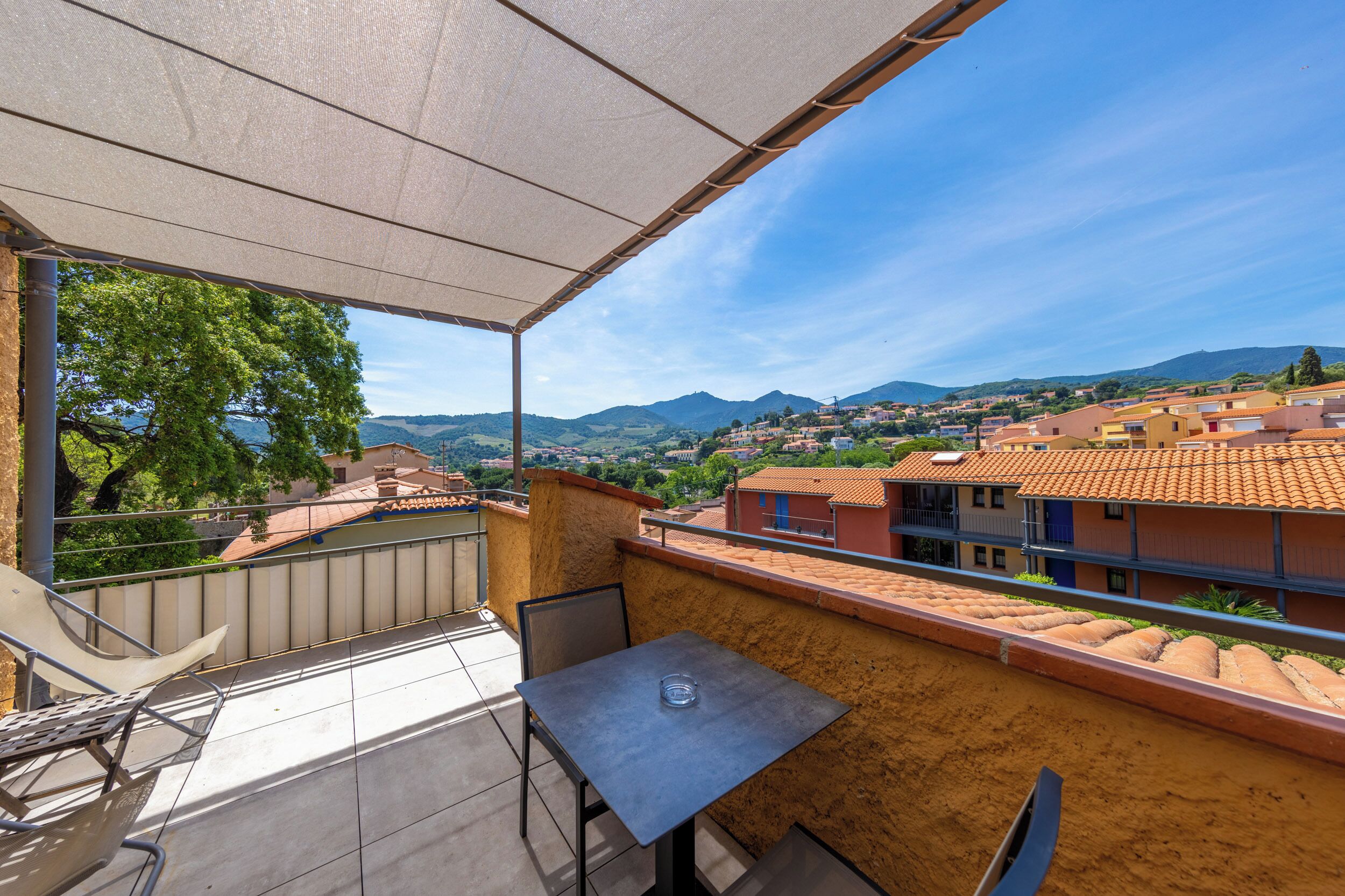 A balcony with a table and chairs, offering a view of a town with terracotta-roofed buildings and distant mountains.