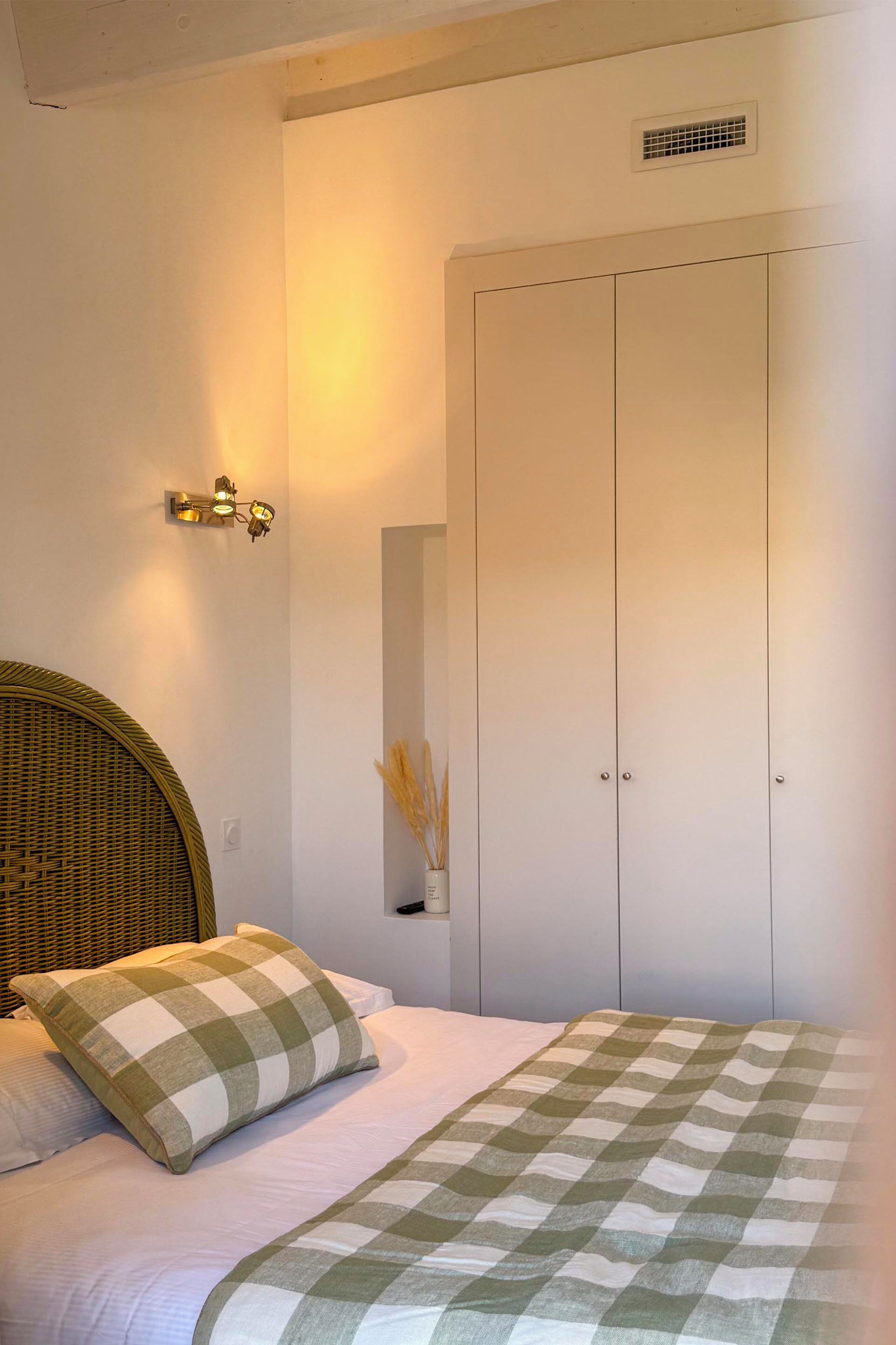 Une chambre à coucher avec un lit, une couverture à carreaux, une tête de lit en osier, un luminaire fixé au mur et une armoire.