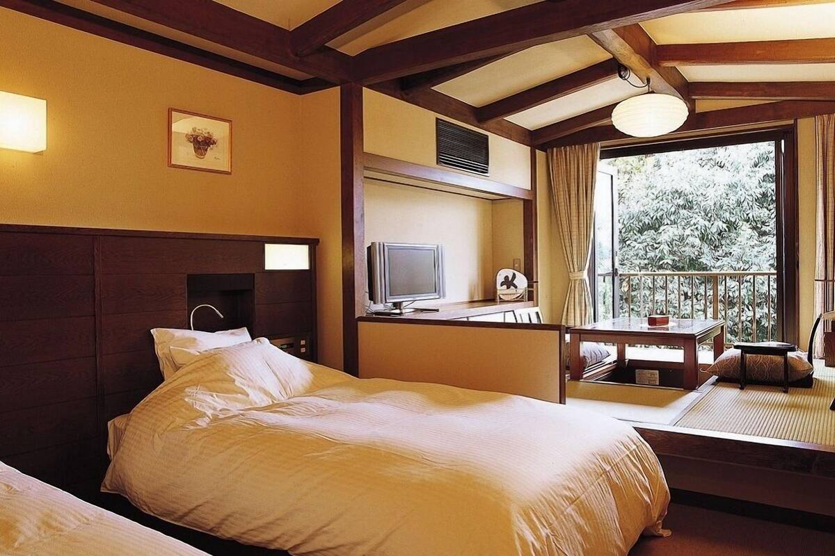A hotel room with a large bed, a desk with a television, and a balcony with a view of trees.