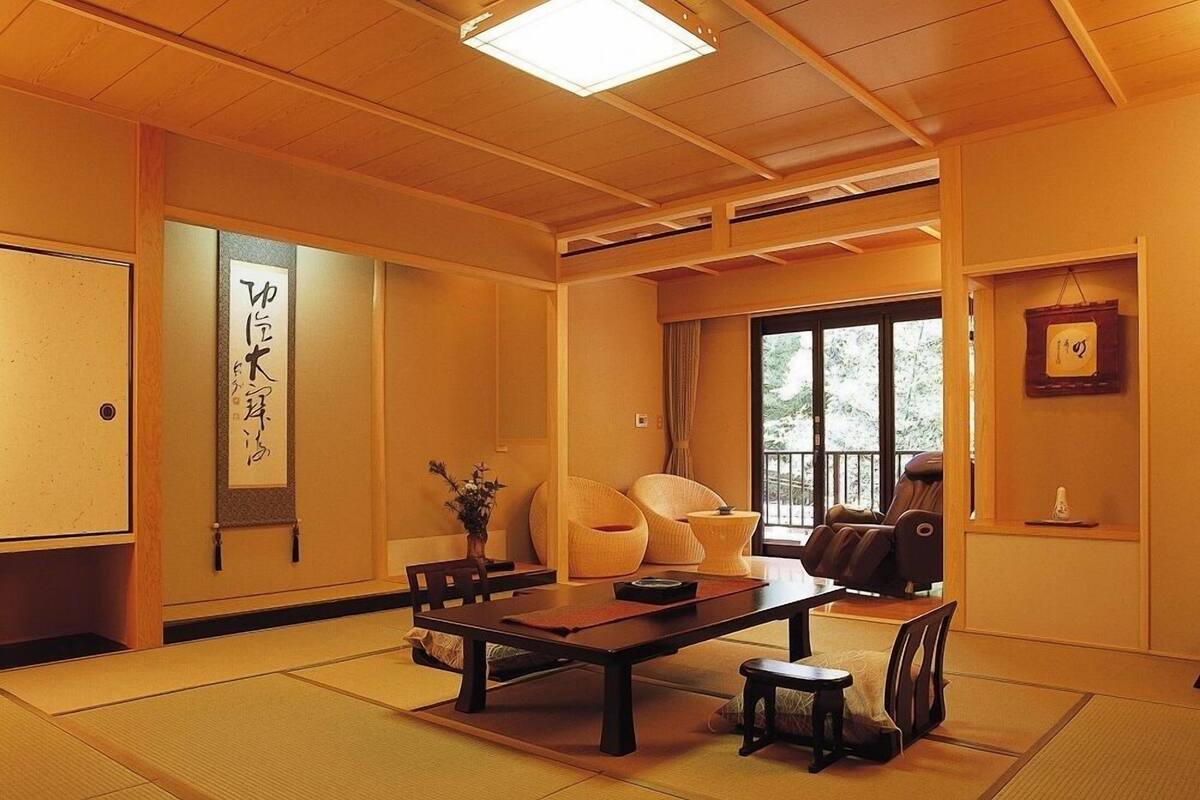 A traditional Japanese room with tatami flooring, a low table, chairs, a massage chair, and a large window.