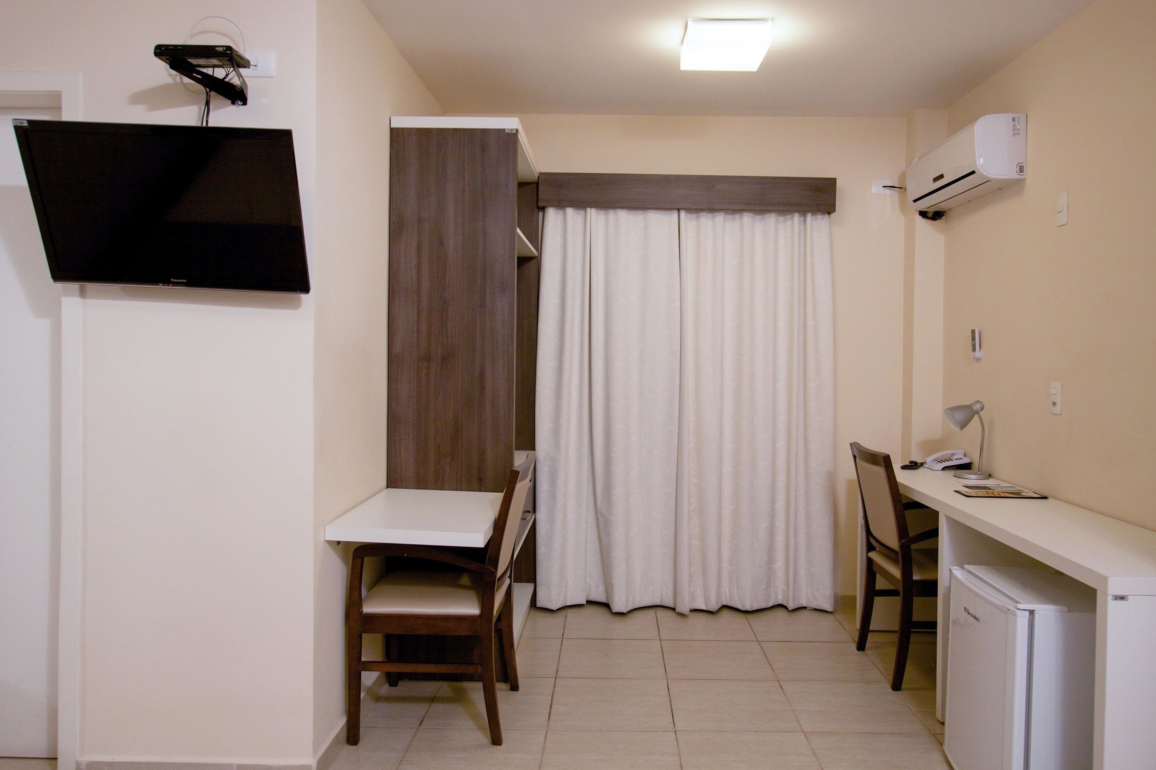 Una habitación pequeña con escritorio, silla, televisor, aire acondicionado y una ventana con cortinas.