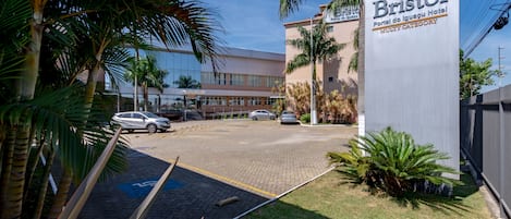 Uma área de estacionamento com um letreiro para o Bristol Hotel, apresentando um edifício moderno com janelas de vidro e palmeiras.