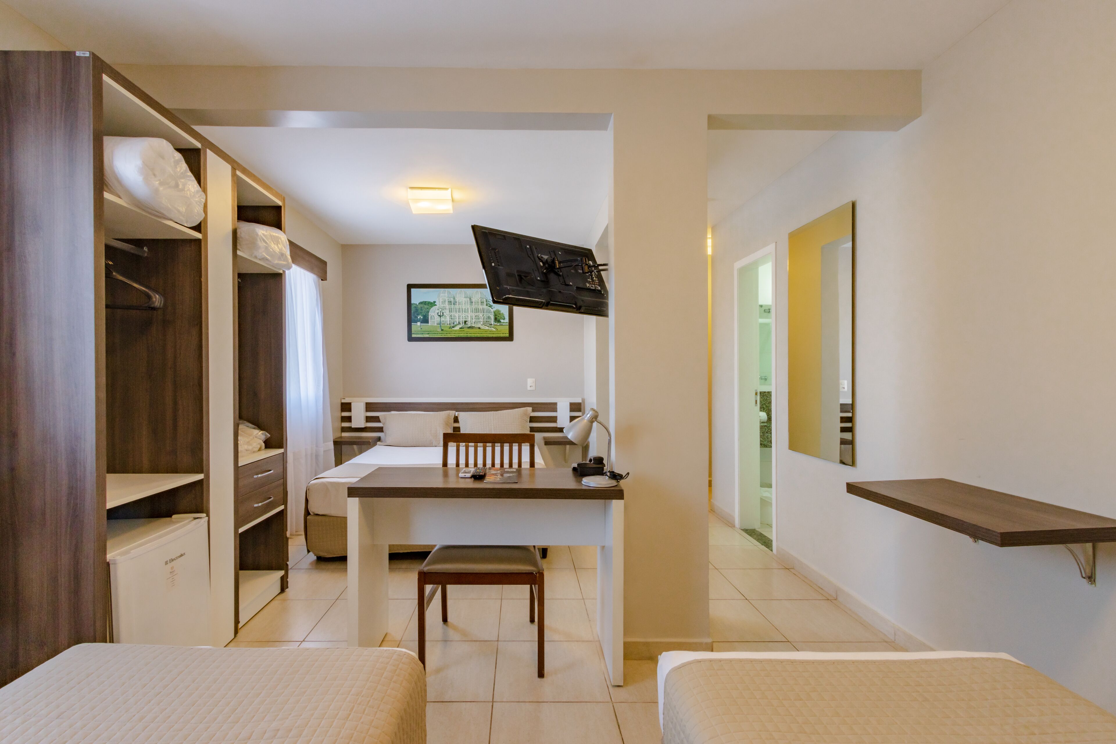 Une chambre d’hôtel comprenant un lit, un bureau avec une chaise, un téléviseur fixé au mur, une armoire et une salle de bain visible à travers une porte ouverte.