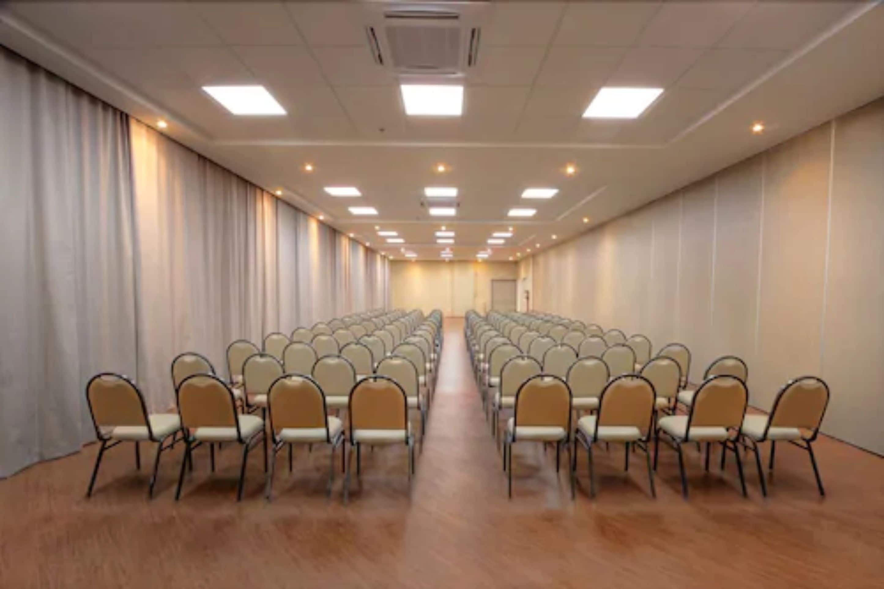 A conference room with rows of chairs and a long table, equipped with ceiling lights and a carpeted floor.