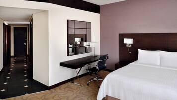 A hotel room with a large bed, a desk with a chair, a mirror, and a wall-mounted lamp.