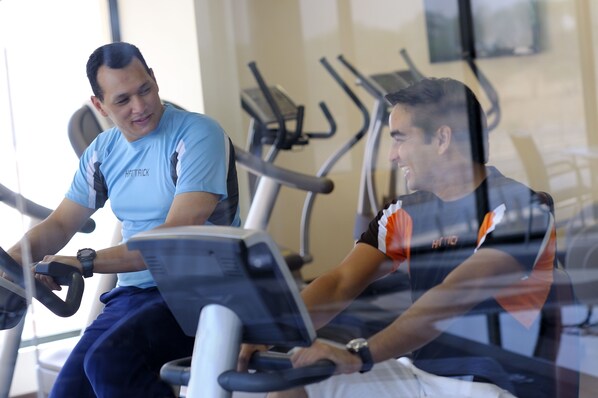 Two individuals on stationary bikes in a gym with exercise equipment in the background.