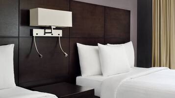 Quarto de hotel com duas camas, uma cabeceira de madeira escura, uma mesinha de cabeceira branca e uma luminária fixada na parede.