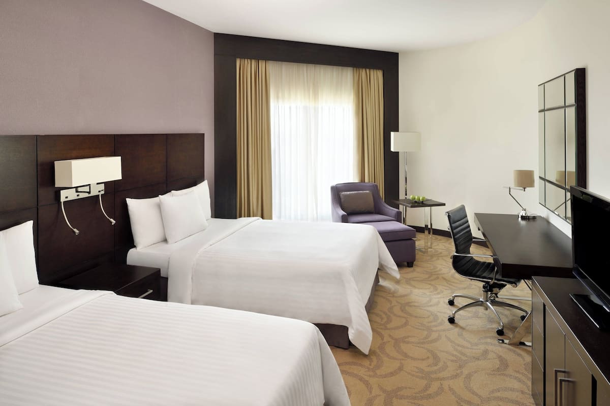 A hotel room with two beds, a desk with a chair, a television, and a large window with curtains.