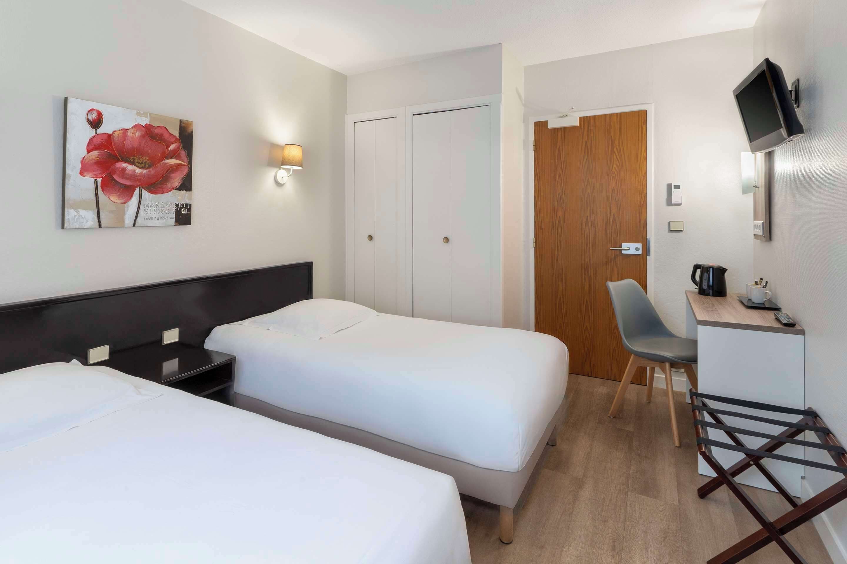 Habitación de hotel con dos camas individuales, un escritorio con silla, un televisor montado en la pared y una puerta de madera.