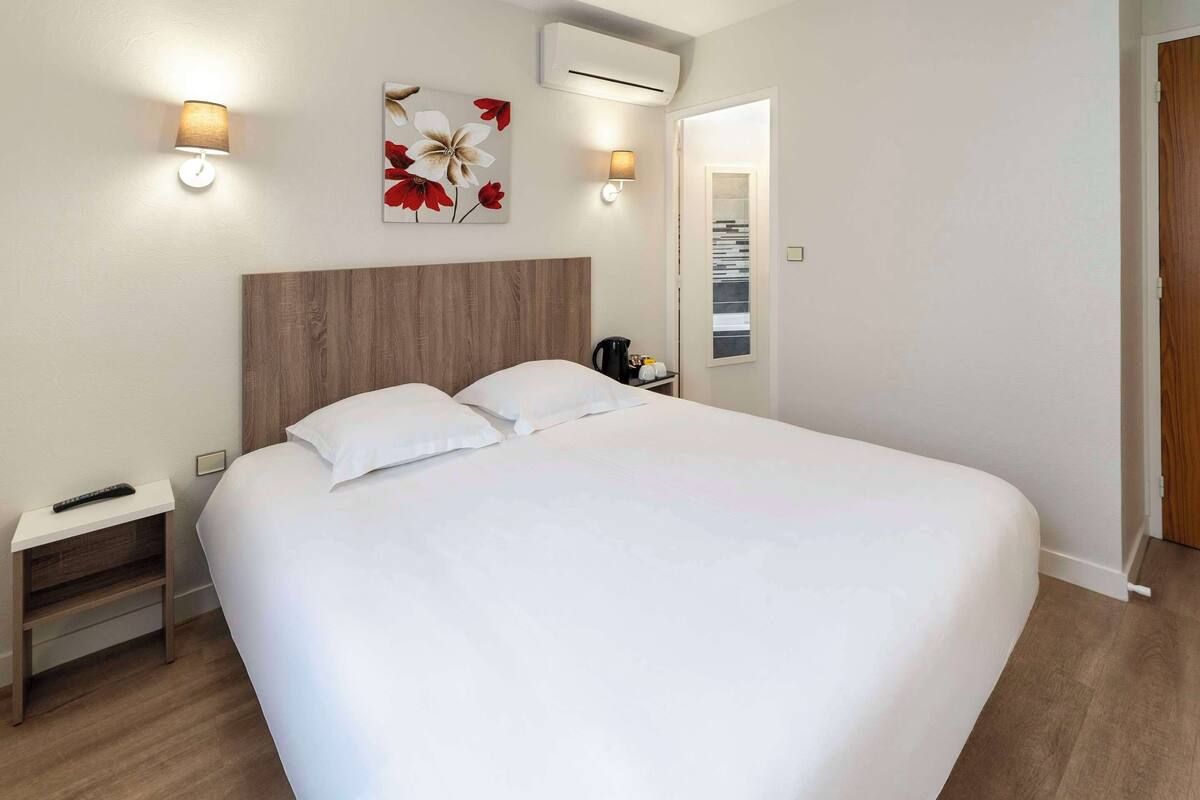 Une chambre d’hôtel avec un grand lit, des tables de chevet, une petite table, une lampe fixée au mur et un tableau.