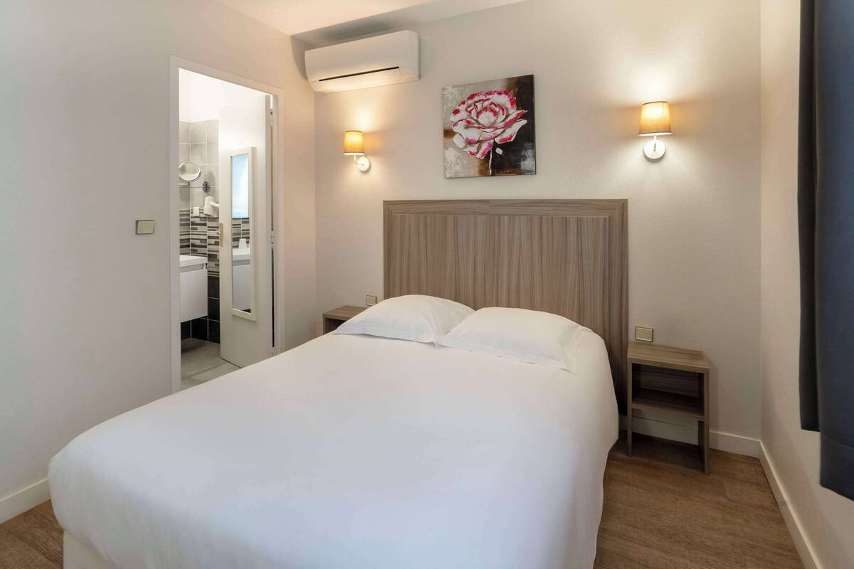 Une chambre à coucher comprenant un lit, deux tables de chevet, une commode, un climatiseur fixé au mur et un tableau représentant une fleur.