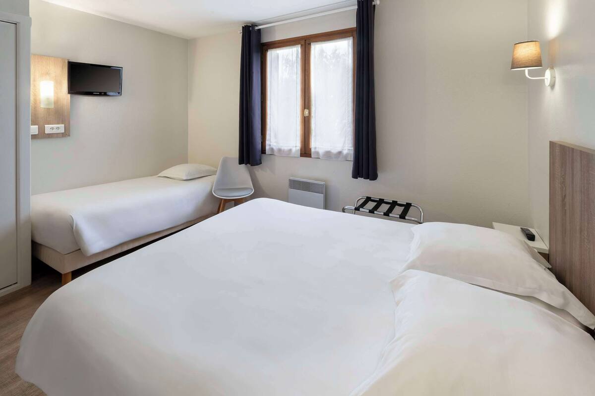 Une chambre d’hôtel avec deux lits, un téléviseur fixé au mur, un radiateur et une fenêtre avec des rideaux.