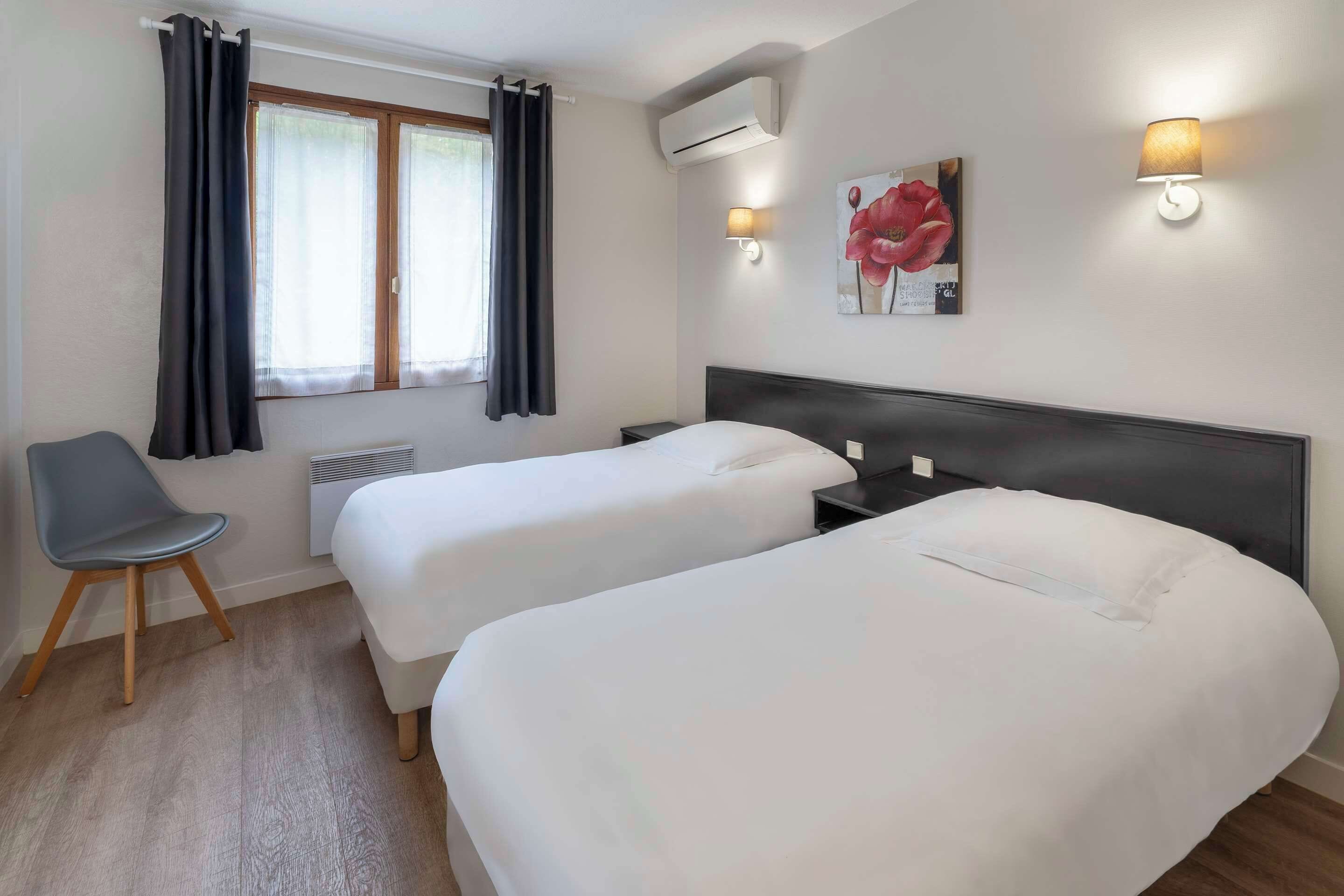 Una habitación de hotel con dos camas individuales, una silla, un ventanal con cortinas, aire acondicionado y una lámpara fijada a la pared.