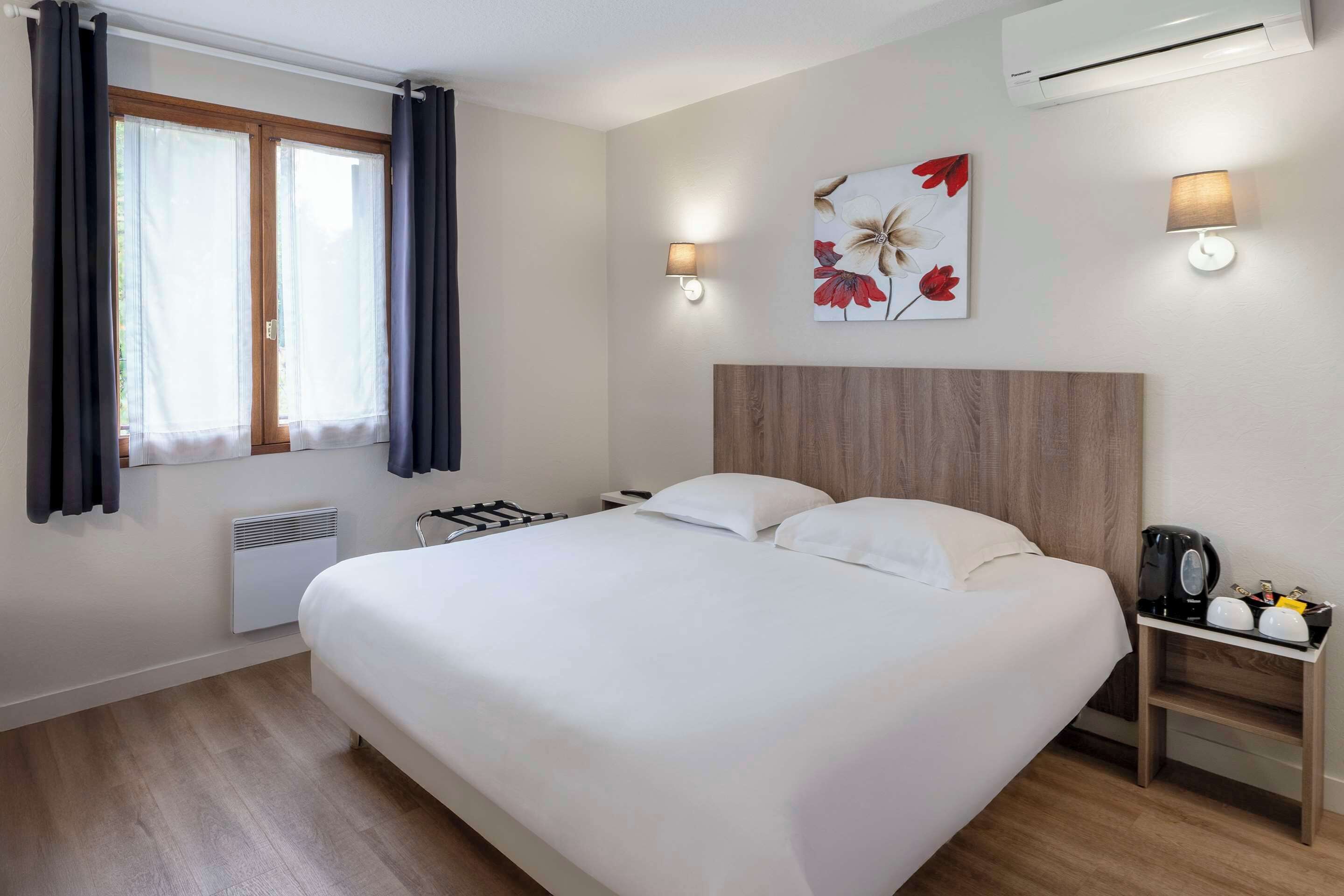Une chambre d’hôtel avec un grand lit, deux tables de chevet, un climatiseur fixé au mur, une fenêtre avec des rideaux et une œuvre d’art florale accrochée au mur.