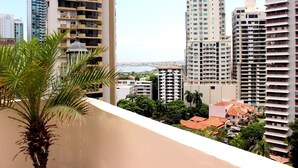 Un edificio de gran altura con varios balcones, una palmera en primer plano y vistas de otros edificios al fondo.