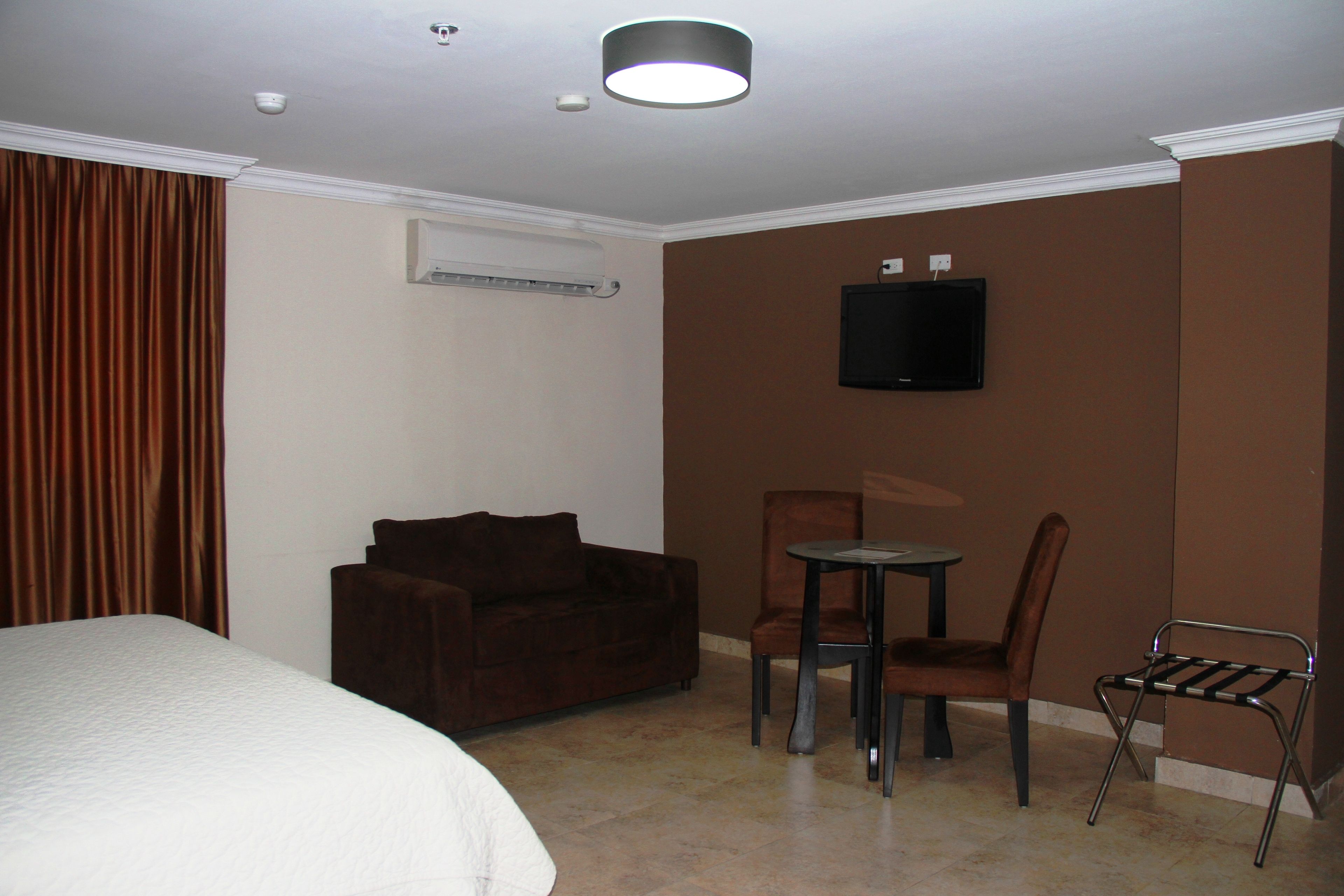 Una habitación de hotel con cama, un sofá marrón, una mesita con una silla y un televisor fijado a la pared.