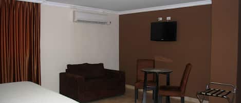 Una habitación de hotel con cama, un sofá marrón, una mesita con una silla y un televisor fijado a la pared.