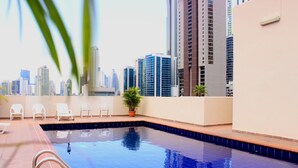 Piscina en la azotea con vistas a un horizonte urbano, rodeada de sillas de descanso y una planta en maceta.