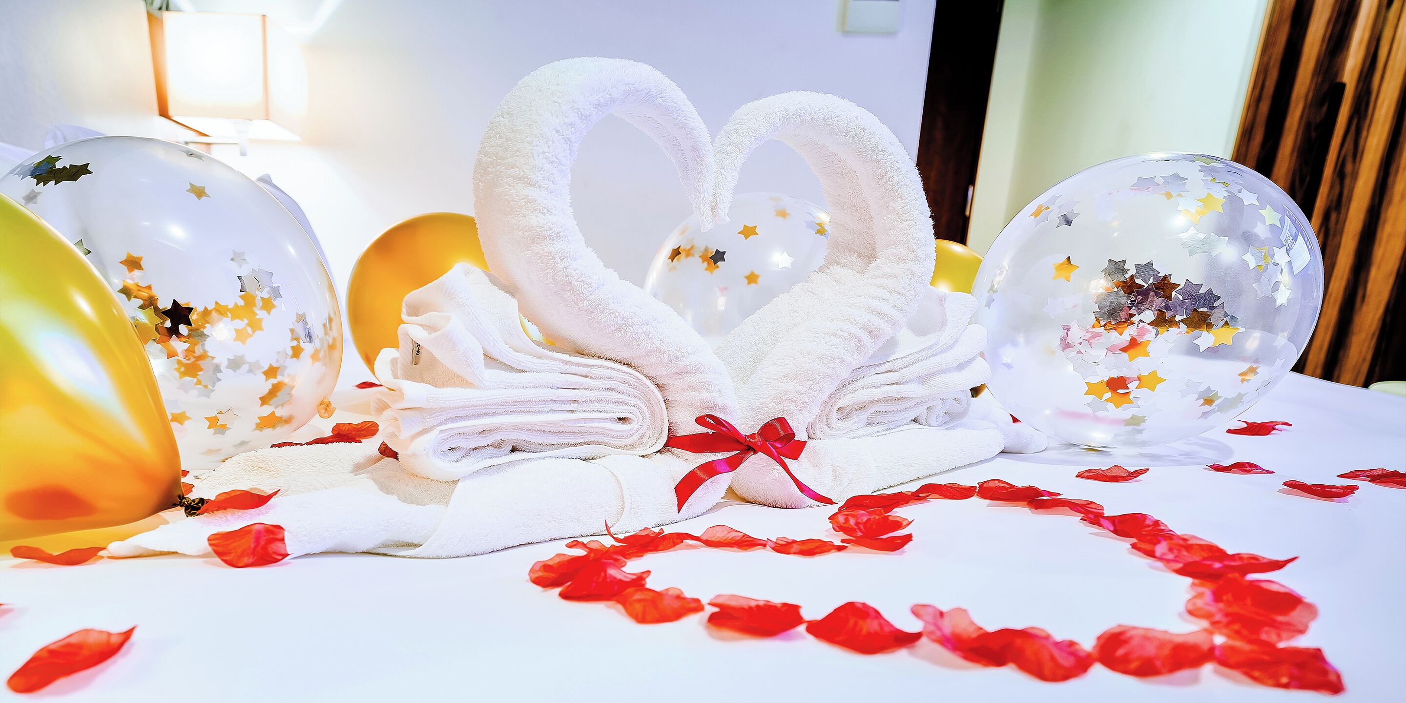 A bed with white towels arranged in a heart shape, surrounded by red rose petals, and two clear balloons with star decorations.