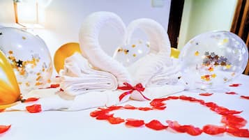 A bed with white towels arranged in a heart shape, surrounded by red rose petals, and two clear balloons with star decorations.