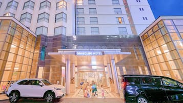 L’extérieur de l’hôtel Mercure se caractérise par une façade moderne en verre, une entrée centrale et deux voitures garées devant.