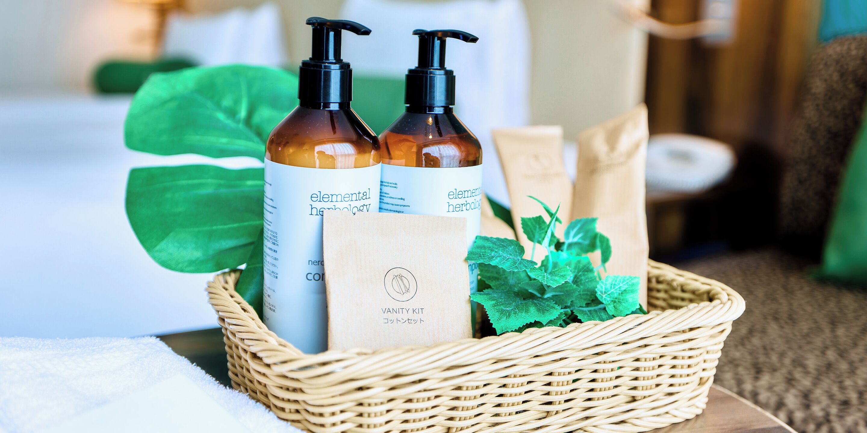 Un panier en osier contenant deux bouteilles de produits de la marque Elemental Herology, un sac en papier et des feuilles vertes.