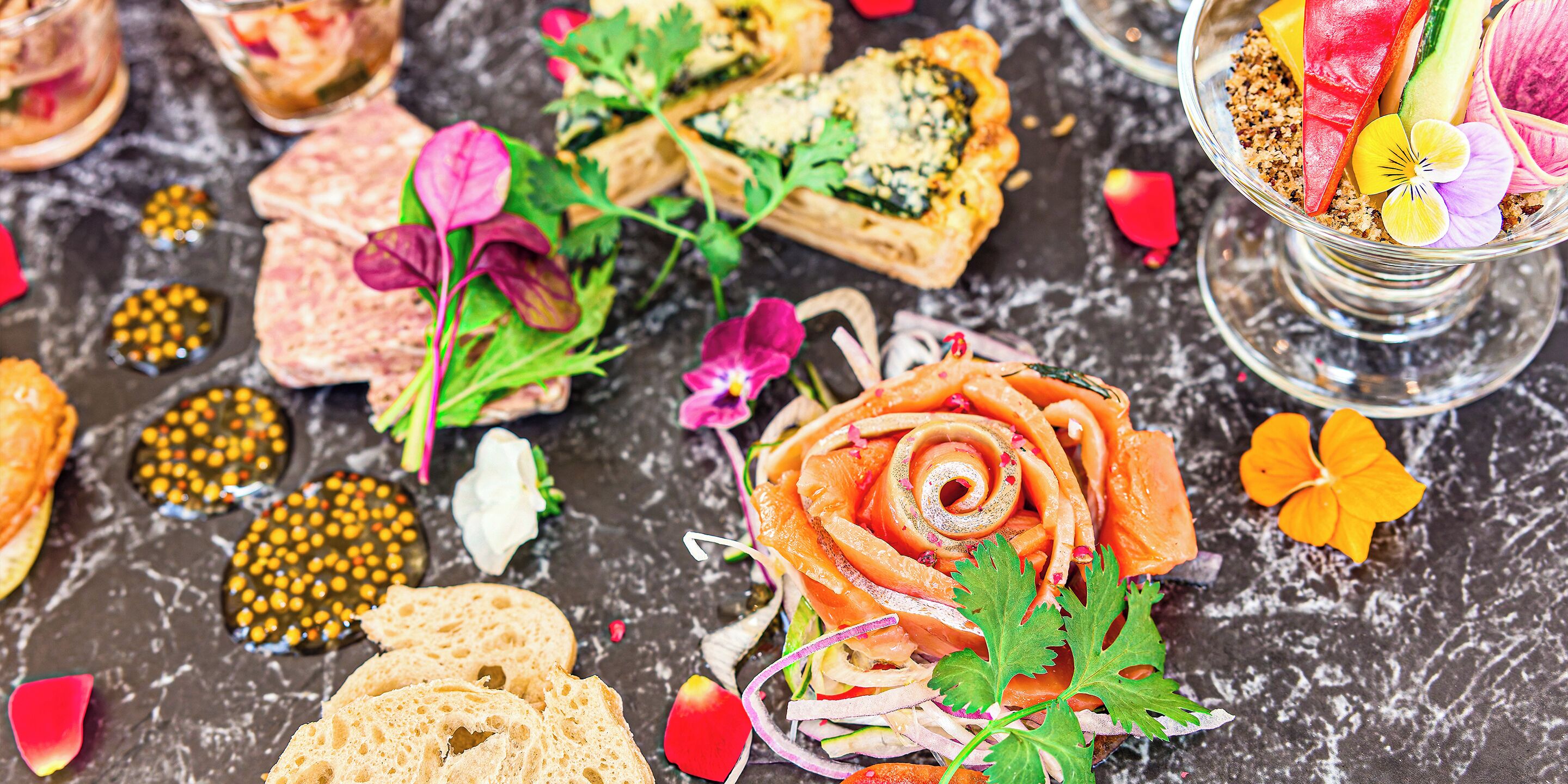 Une variété d’aliments sont présentés, notamment un plat de saumon disposé en forme de rose, du pain et diverses garnitures, le tout sur une surface en marbre.
